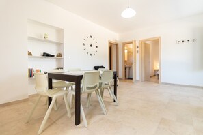 A dining area with a table, chairs, a wall clock, shelves with books, and a door leading to another room.