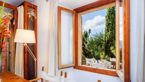 A room with a wooden table, a hanging chair, a lamp, and a window with a view of greenery and a table outside. - Vacation Home "Casa en Centro Histórico Pollença" with Private Terrace, Garden & Wi-Fi (pollensa)