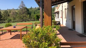 Un patio al aire libre techado con una mesa y bancos de madera, rodeado de follaje y una vista de una casa al fondo.