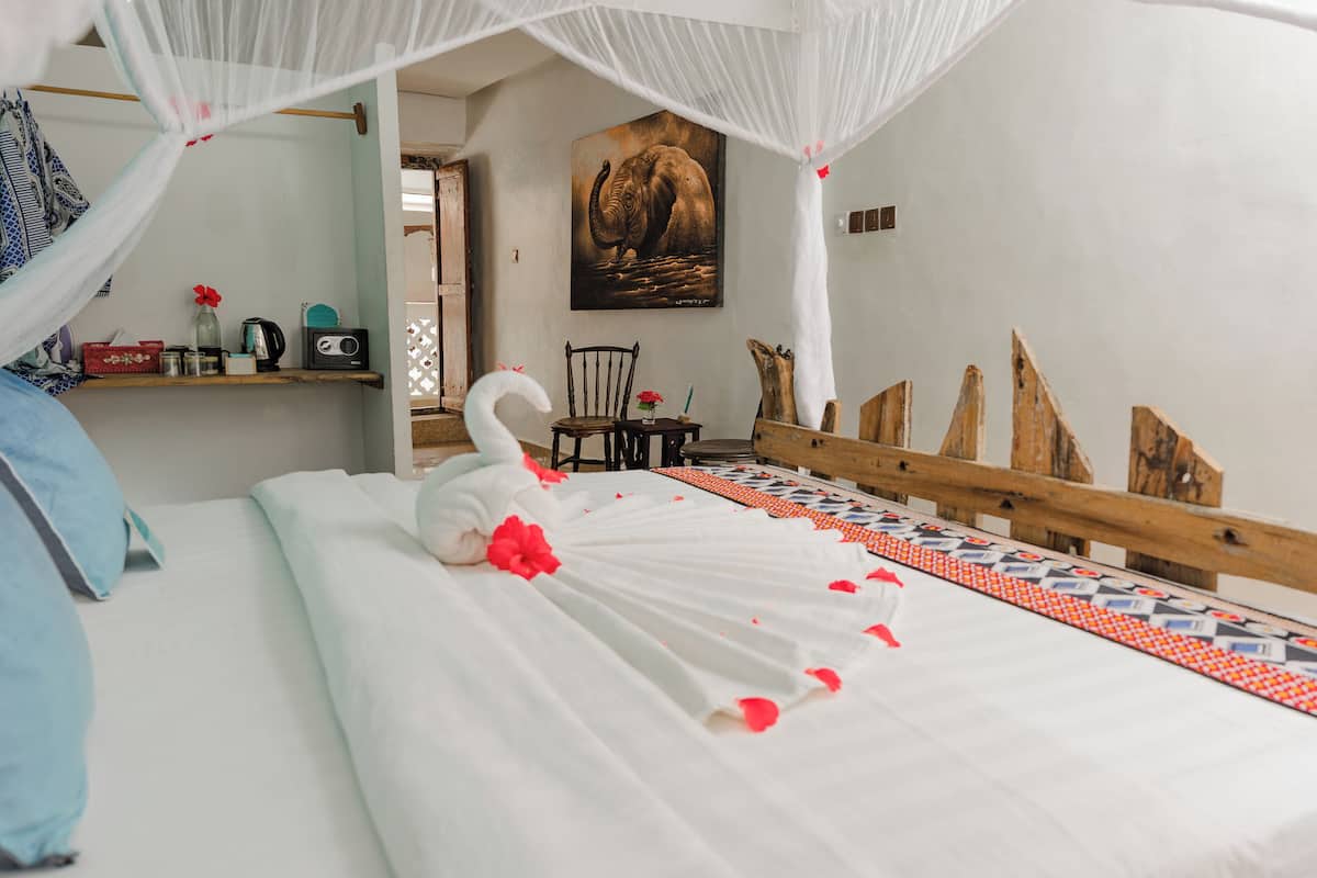 Una cama con dosel, cabecera de madera, un cuadro de un elefante y una mesita con un jarrón y flores.