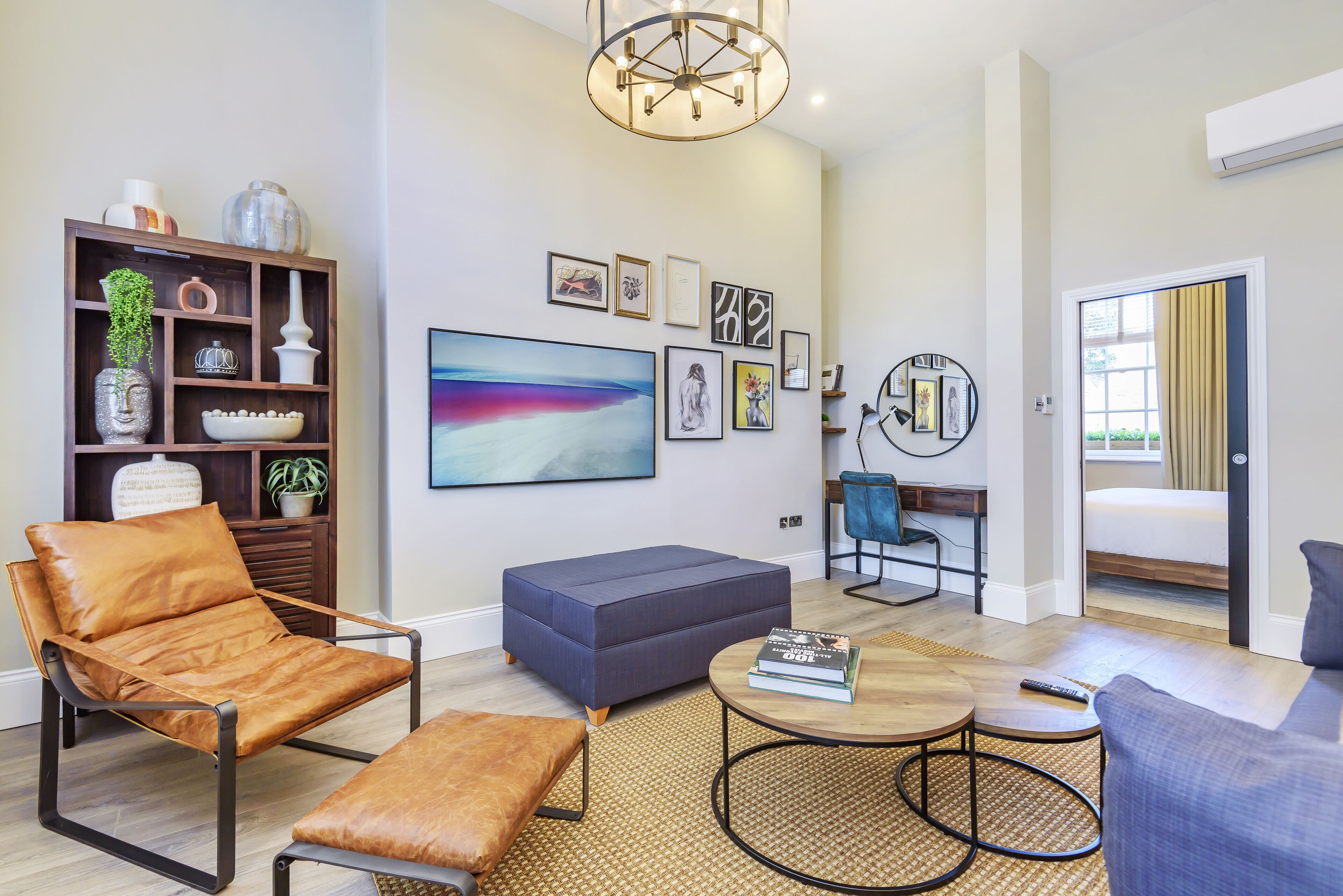 Een moderne woonkamer met een leren fauteuil, een blauwe bank, een ronde salontafel en een boekenplank met decoratieve objecten.