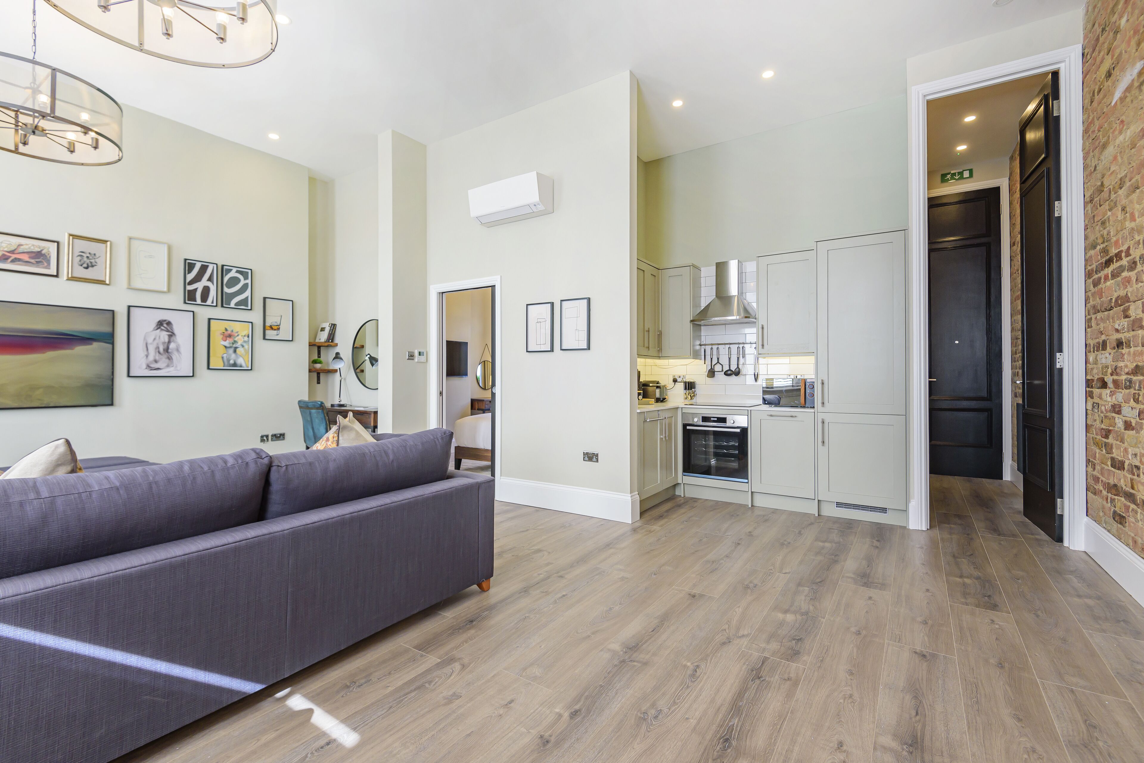 Een moderne woonkamer met een grijze bank, een keuken met witte kastjes en een bakstenen muur.