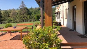 En overdækket terrasse med et træbord og bænke, omgivet af grøn vegetation og udsigt til et hus i baggrunden.