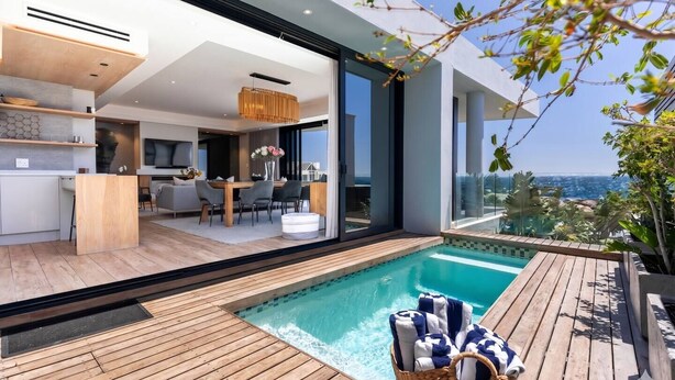 Piscina moderna ao ar livre com deck de madeira, área de refeições com cadeiras e mesa, e vista para o oceano.