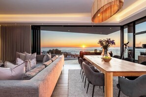 A modern living room with a large window overlooking the ocean at sunset, a wooden dining table, and a comfortable seating area. - Little Beach Villa-200m from the beach, best views (Cape Town)