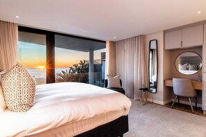 Une chambre d’hôtel avec un grand lit, un bureau, une chaise, un miroir rond et une vue sur le coucher de soleil sur l’océan.