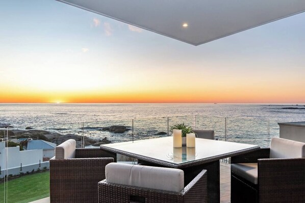 An outdoor seating area with a view of the ocean at sunset, featuring a table and wicker chairs. - Little Beach Villa-200m from the beach, best views (Cape Town)