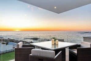 An outdoor seating area with a view of the ocean at sunset, featuring a table and wicker chairs.