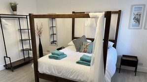 Ein Himmelbett mit weißer Bettdecke und türkisfarbenen Handtüchern, ein Nachttisch, ein Wandregal und ein Blumentopf.
