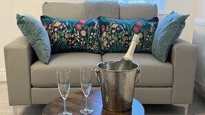 Ein Wohnzimmer mit einem beigen Sofa, floralen Kissen, einem Metallkübel mit einer Flasche und zwei Gläsern auf einem Holztisch.