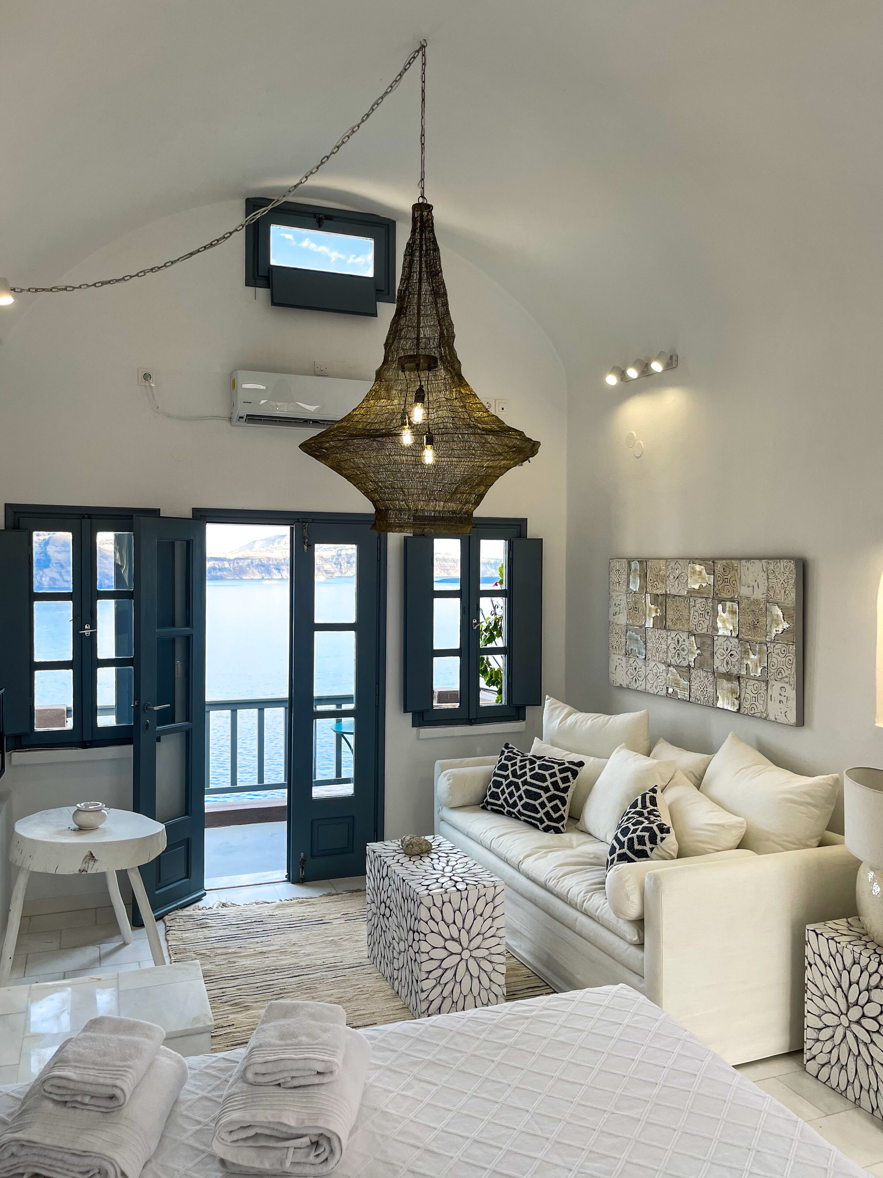 A modern bedroom with a large bed, a white sofa, a patterned coffee table, and a view of the sea through open doors.