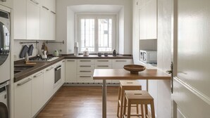 A modern kitchen with white cabinets, a wooden countertop, a large window, and a dining area with stools.
