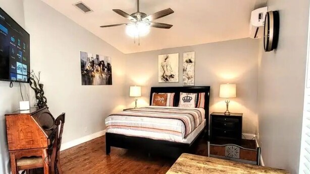 A bedroom with a bed, bedside tables, a ceiling fan, wall art, and a TV mounted on the wall.
