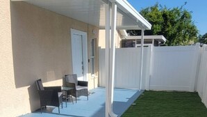 A covered patio area with a white ceiling, a small seating area with chairs and a table, and a green artificial lawn.