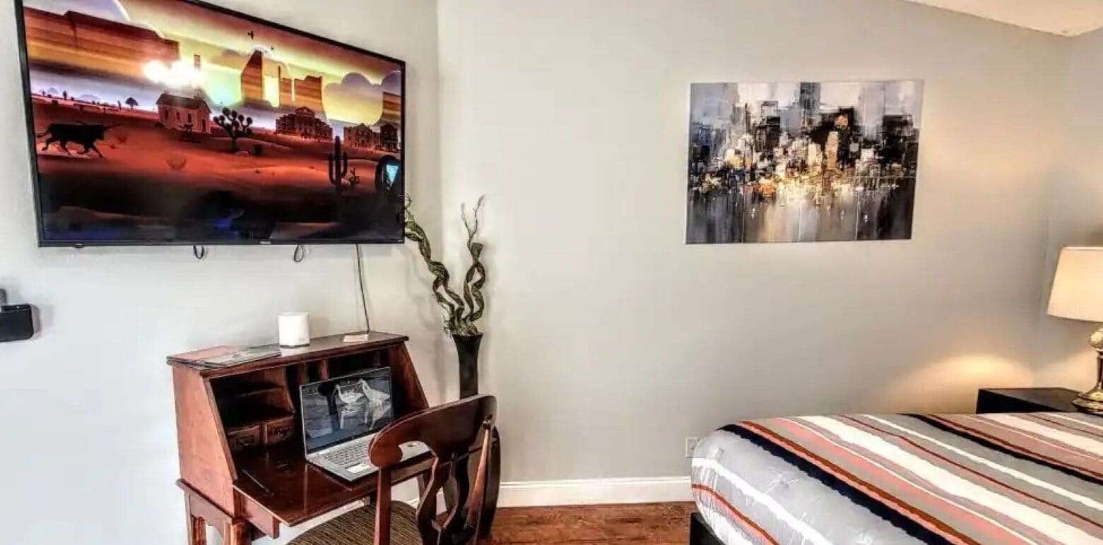 Habitación de hotel con cama, escritorio con una computadora portátil, televisor en la pared y dos cuadros.