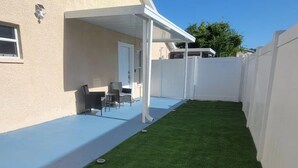 A covered patio area with a small lawn, white walls, and a white fence.