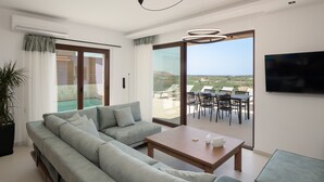 A modern living room with a sofa, coffee table, and a TV. There is a sliding glass door leading to an outdoor patio with a table and chairs. - Anema Villa, a picture-perfect summer escape (Rethymno)