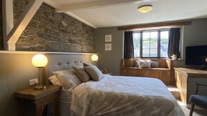 A bedroom with a stone wall, a bed with a headboard, bedside lamps, a window with a view, and a desk with a chair. - Tom's of Tregaron (Tregaron)