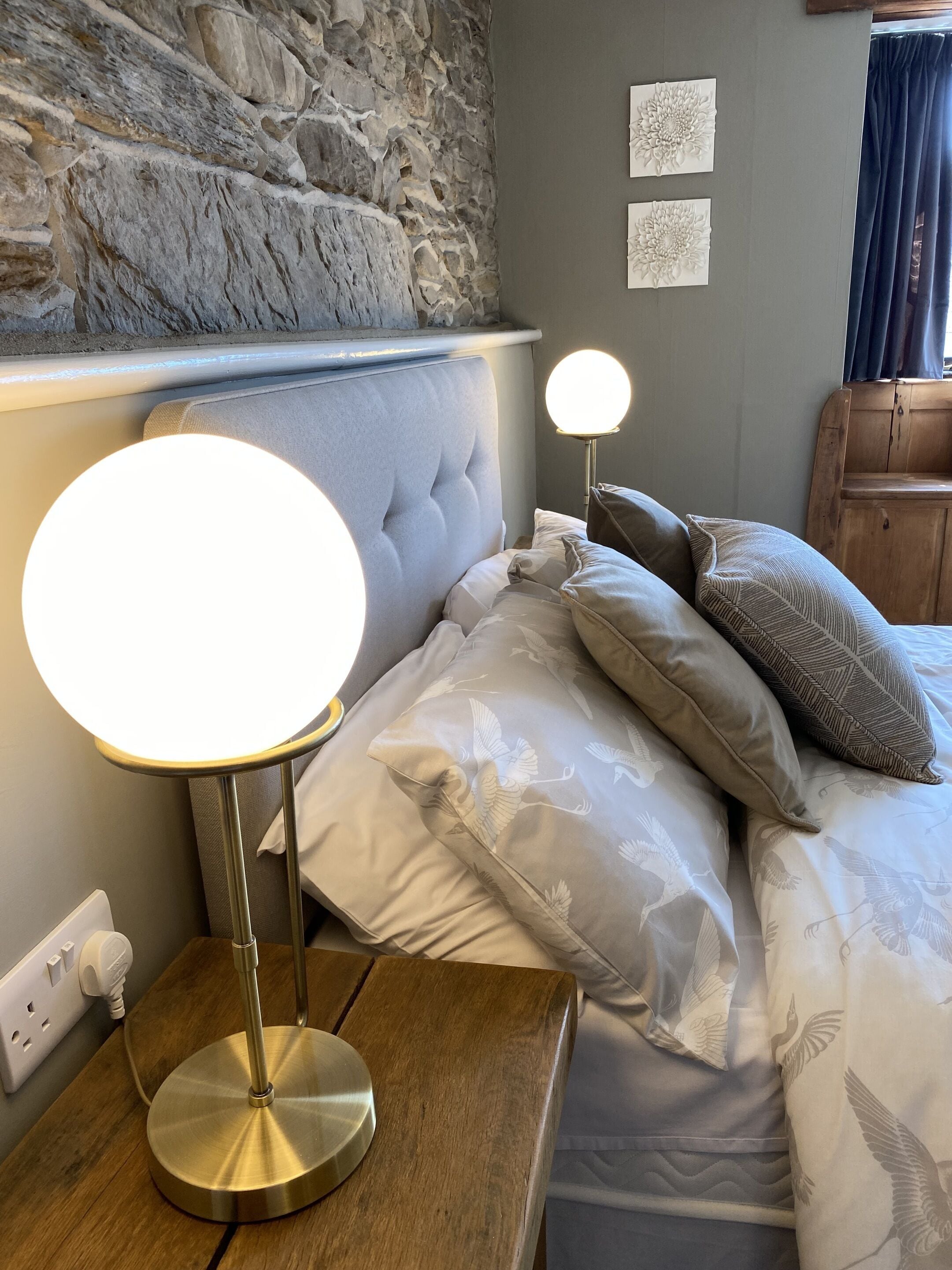 A neatly made bed with white bedding and patterned pillows, a round table lamp, a stone wall, and a wooden nightstand.
