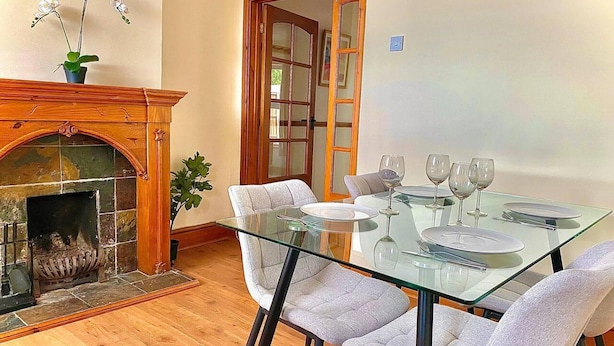 Comedor con una mesa de vidrio y cuatro sillas, una chimenea con una planta, y una puerta con un espejo.