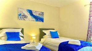 Habitación de hotel con dos camas, cada una con ropa de cama azul y almohadas blancas, un cuadro en la pared y una lámpara en la mesita de noche.