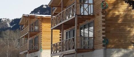 A wooden chalet with multiple balconies and large windows, set against a snowy landscape with mountains in the background.