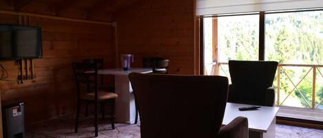 A cozy cabin interior with a wooden ceiling, a dining table with chairs, a TV mounted on the wall, and a window with a view of greenery.