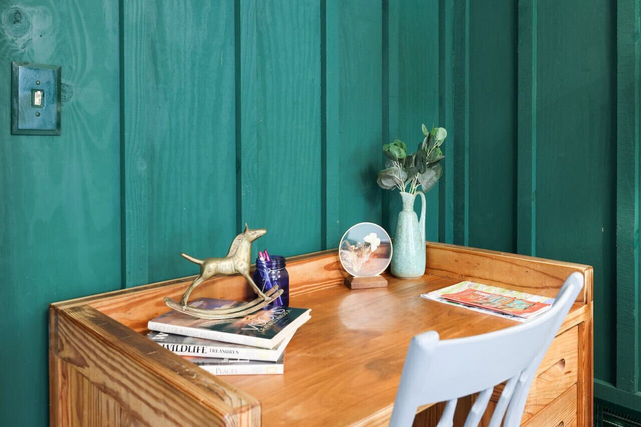 Une table en bois, sur laquelle on trouve des livres, un globe terrestre et un vase avec des plantes, le tout placé devant un mur de couleur turquoise.