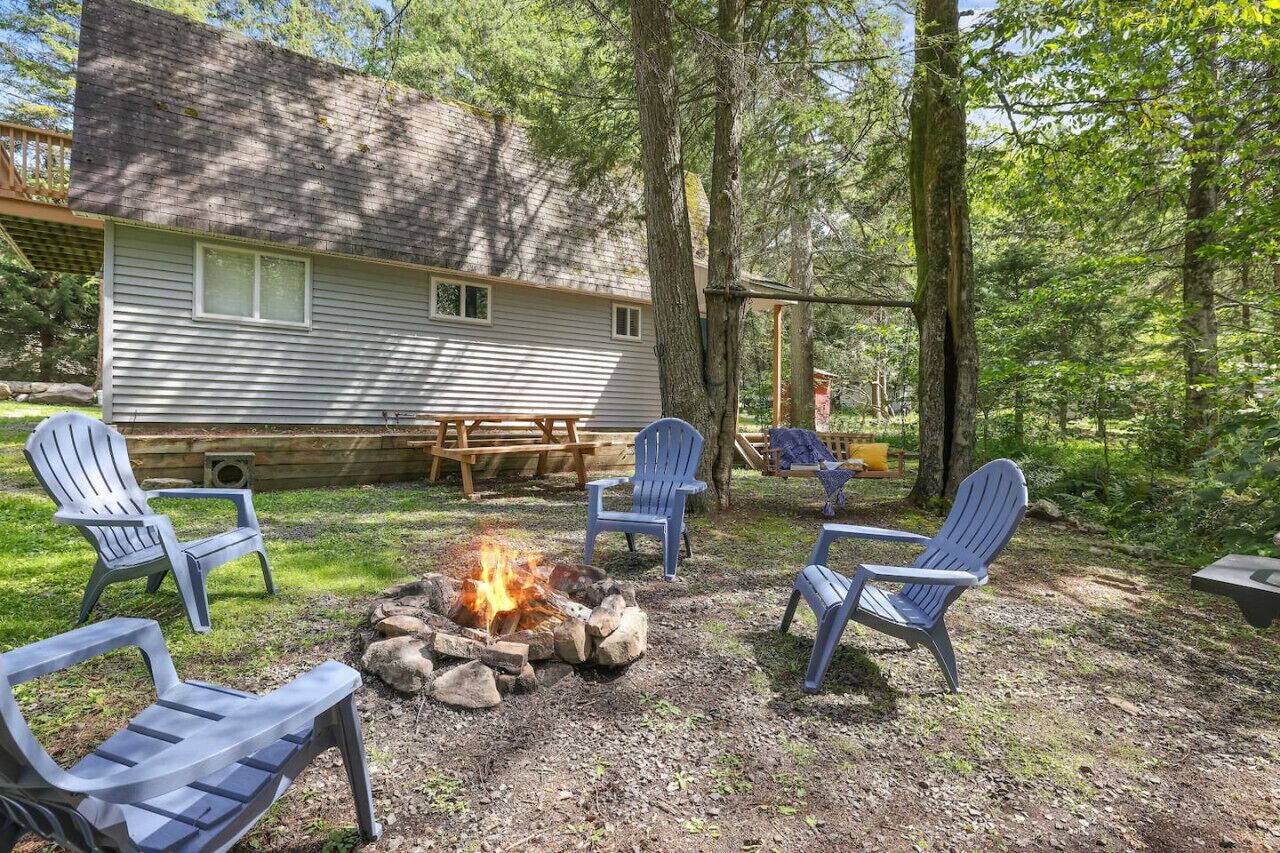 A cabin with a fire pit surrounded by Adirondack chairs, a picnic table, and a covered seating area.