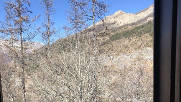 Un balcon doté d’une balustrade en métal, donnant sur un paysage montagneux parsemé de quelques arbres.