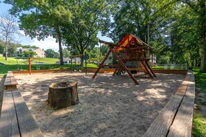 Children can play on the swing set and in the sandpit, surrounded by a fence and shaded by trees.