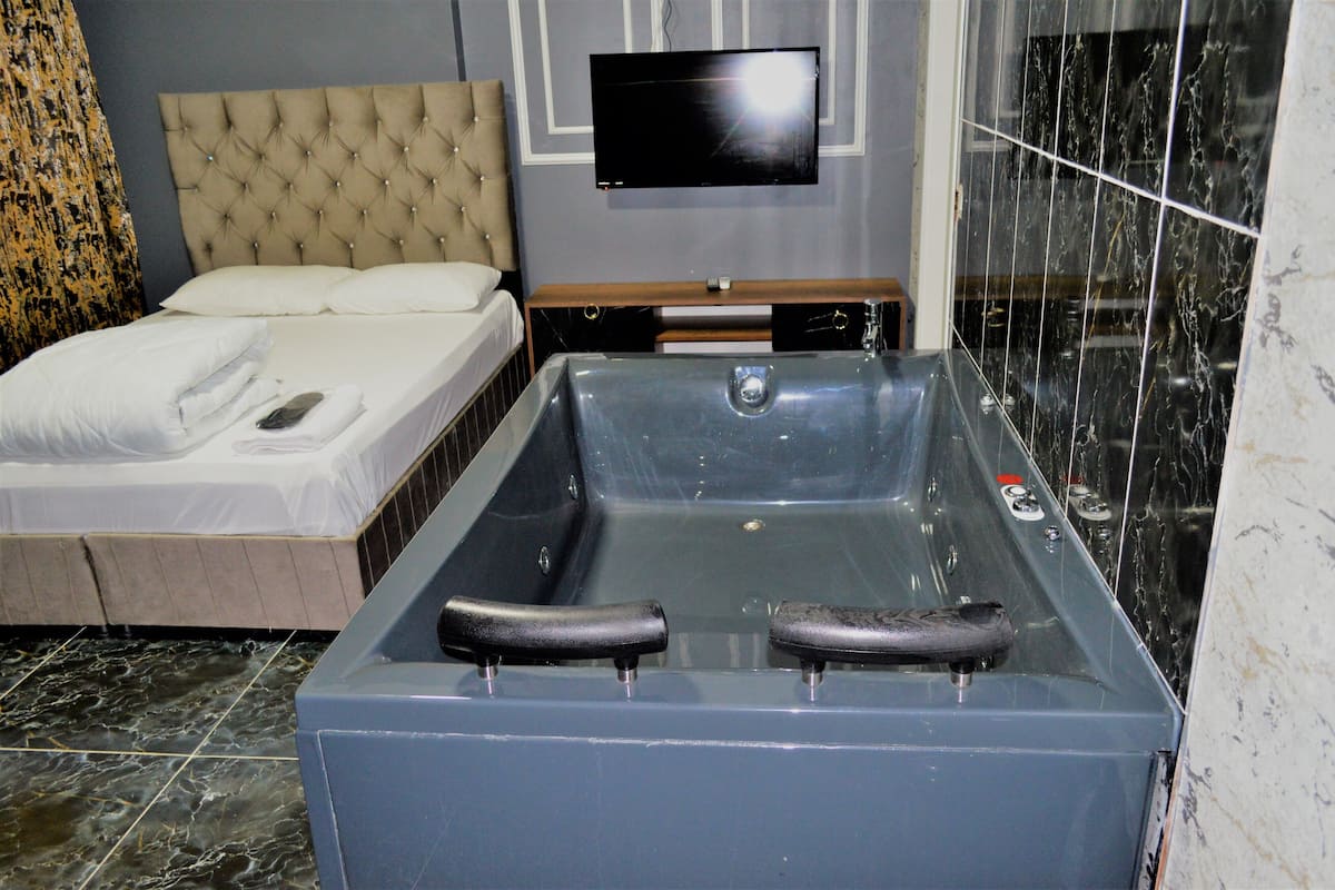 Une salle de bain moderne avec une grande baignoire, un lit recouvert de linge de maison blanc et un téléviseur à écran plat fixé au mur.