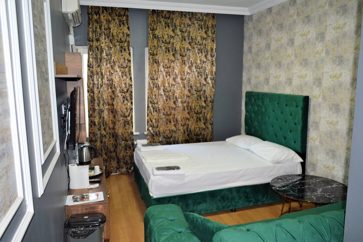 Une chambre d’hôtel comprenant un lit, un banc recouvert d’un tissu vert, une petite table ronde et un espace douche séparé par des rideaux.