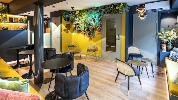 Un intérieur moderne avec un espace salon comprenant des tables et des chaises rondes, un mur décoratif agrémenté de plantes et un mur d’accent jaune.