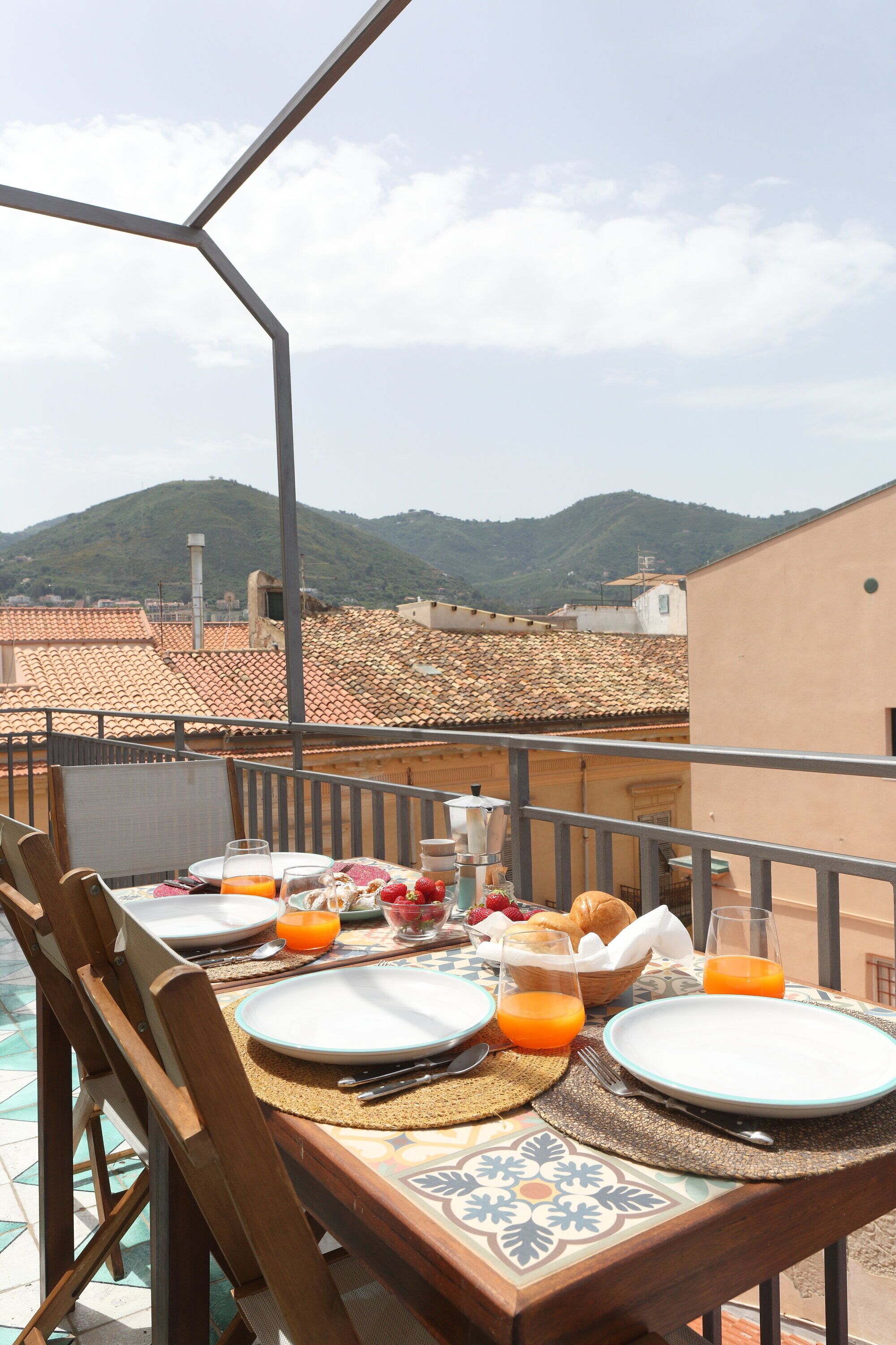 A table set for breakfast with plates, cutlery, and orange juice on a balcony with a view of mountains and terracotta roofs.