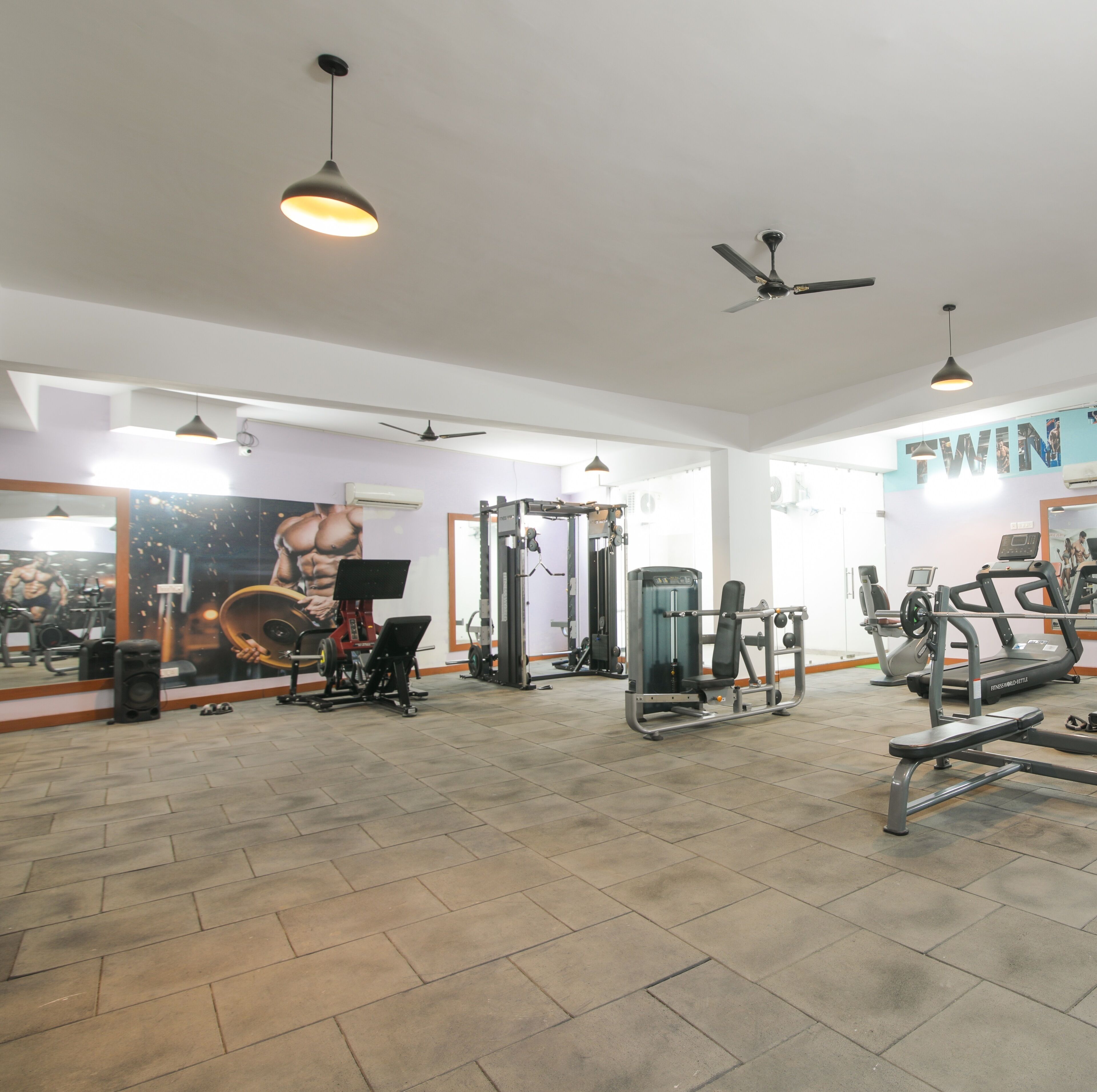 Een fitnessruimte met diverse fitnessapparaten, waaronder een loopband, krachttoestellen en een spiegelwand met fitnessposters.
