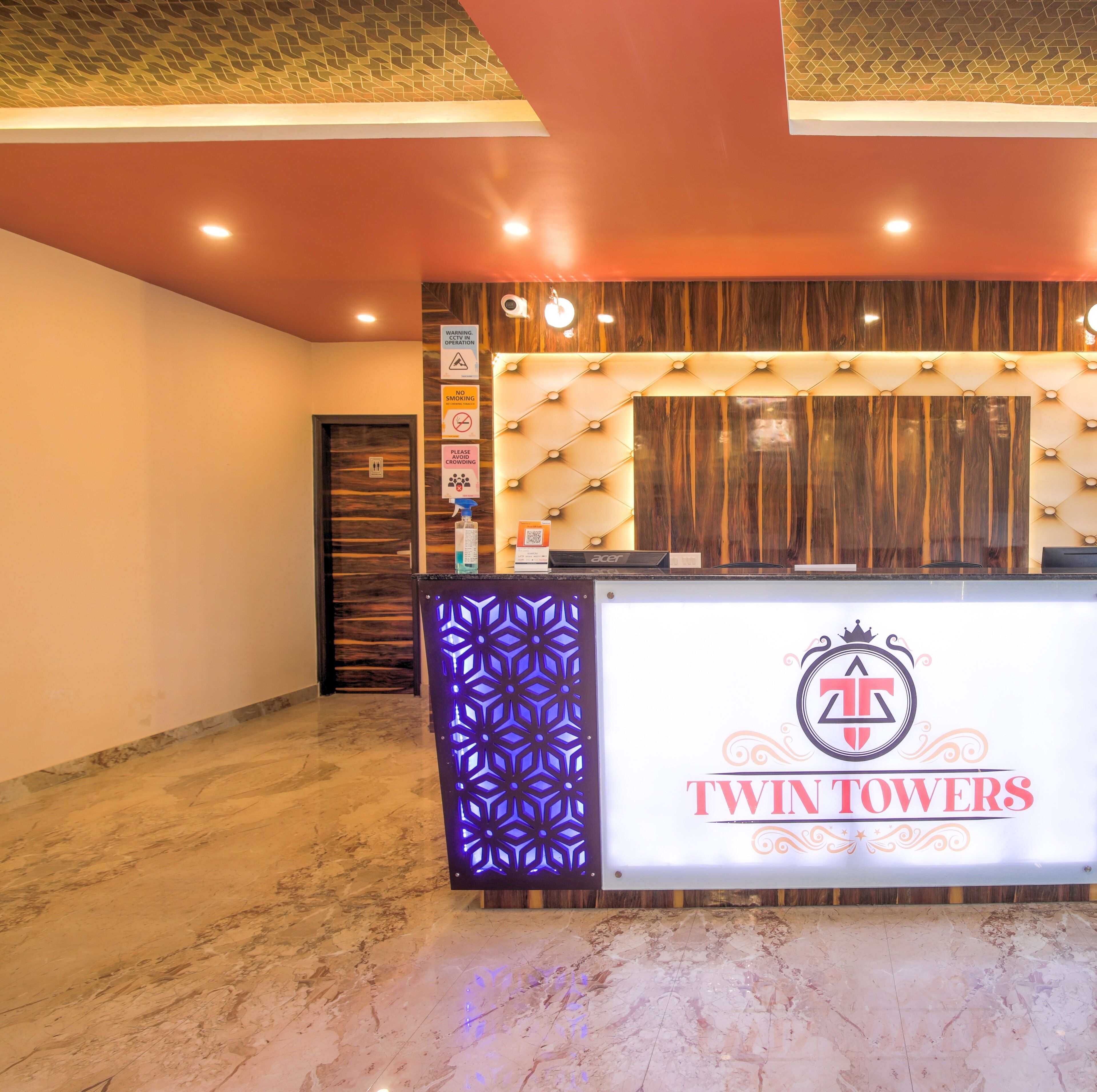 A reception desk with a sign for 'Twin Towers' hotel, a CCTV warning sign, a no smoking sign, and a no crowding sign.