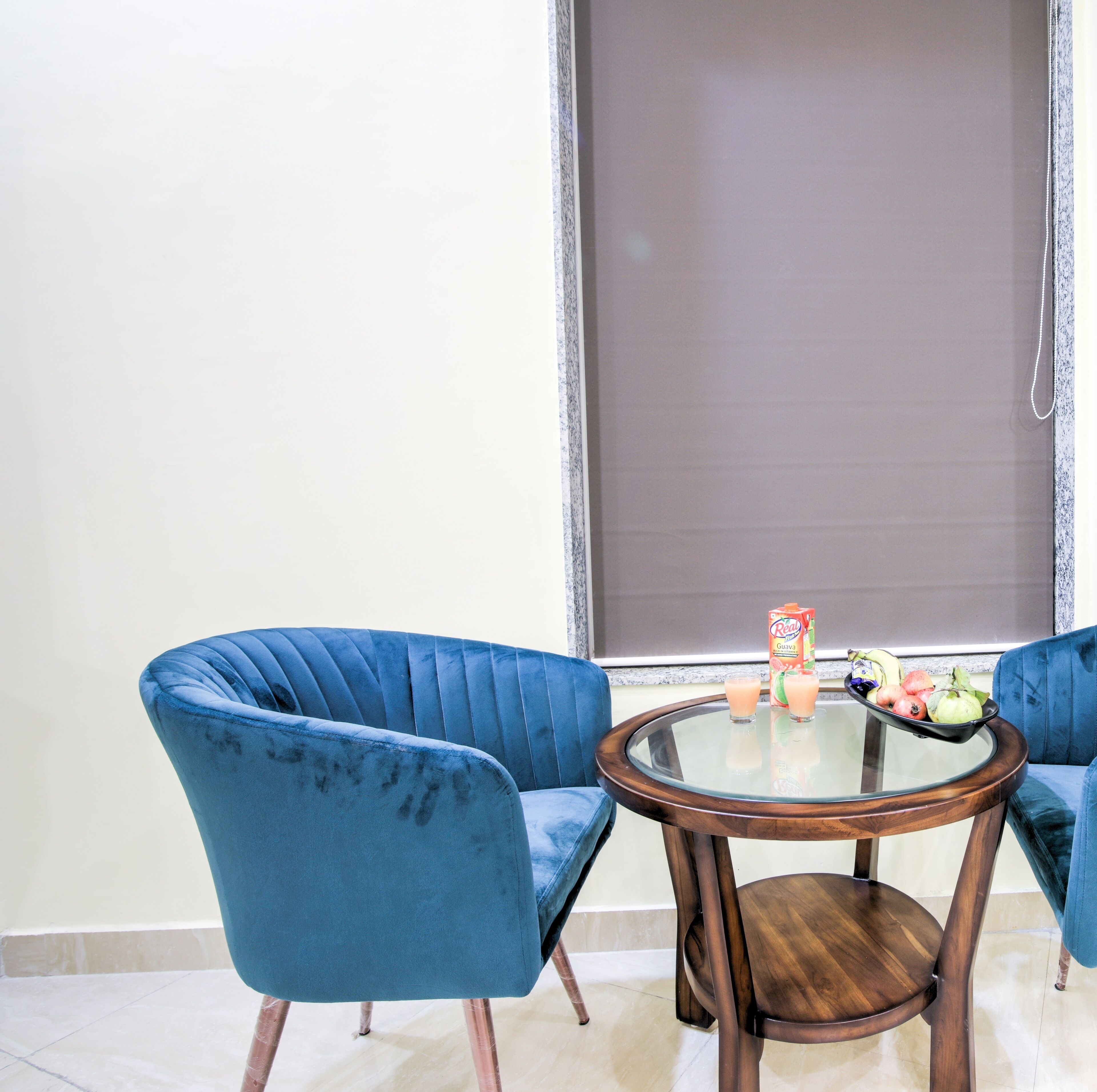 Ein Zimmer mit zwei blauen Sesseln und einem runden Holztisch mit Glasplatte. Ein Fenster mit heruntergelassener Jalousie, und eine Packung Real Guava Saft steht auf dem Tisch.