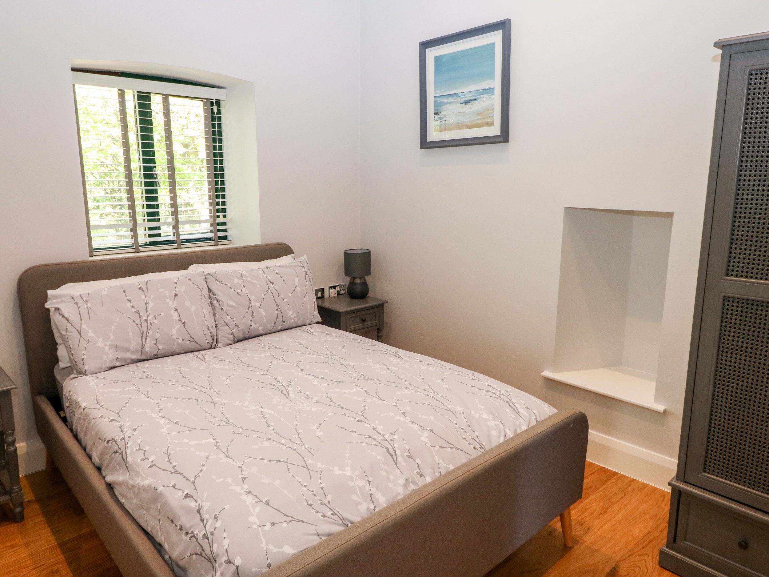 A bedroom with a bed, a nightstand, a lamp, a window with blinds, a framed picture on the wall, and a built-in shelving unit.