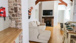 A cozy living room with a stone wall, a beige sofa, a wooden dining table with chairs, and a fireplace. - Ramsey Cottage (Haverfordwest)