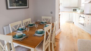 A dining area with a wooden table set for six, white chairs, and teal dinnerware. A framed picture hangs on the wall. - Ramsey Cottage (Haverfordwest)