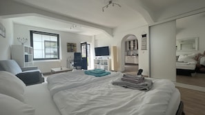 Ett modernt hotellrum med en stor säng, ett skrivbord, en stol, en tv och ett badrum som syns genom en valvöppning.