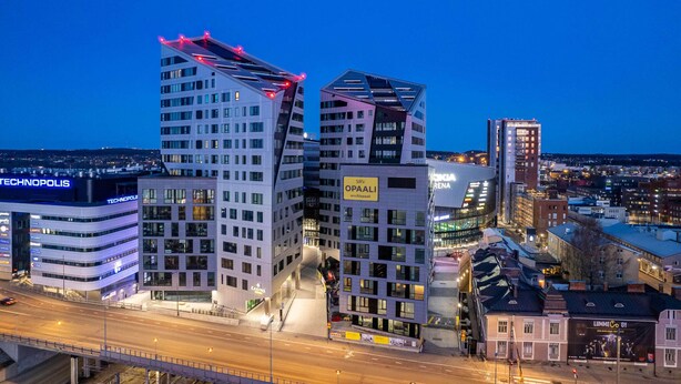 Un paisaje urbano con edificios modernos, incluyendo un edificio destacado con un diseño angular distintivo y un cartel que dice 'SRI OPAALI'.