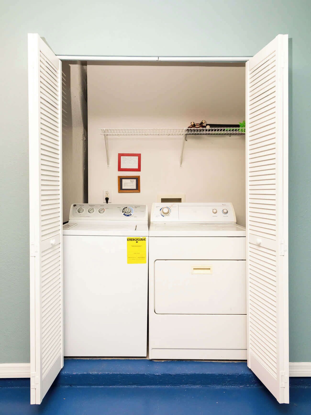 A laundry room with two white washers and dryers, a metal shelf with a red frame, and a yellow energy guide sticker on one of the dryers.