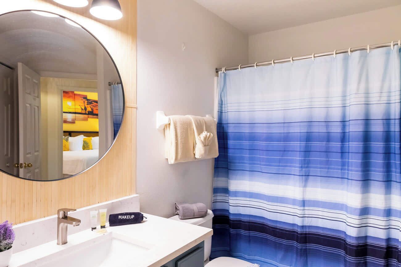 A bathroom with a round mirror, a white sink with a makeup kit, a blue-striped shower curtain, and a towel rack with towels.