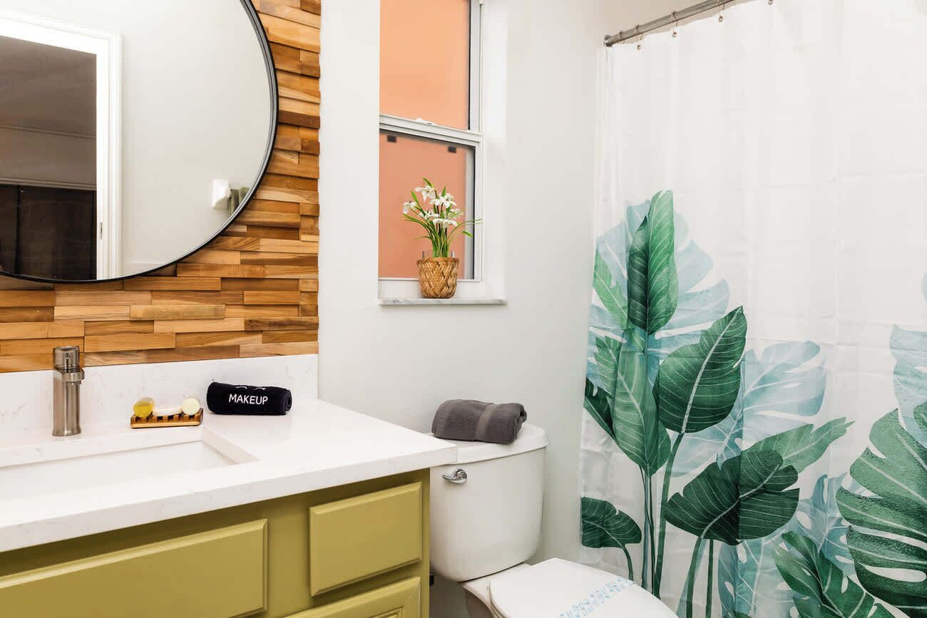 Une salle de bain avec un mur en bois, un miroir rond, un lavabo avec des articles de toilette, des toilettes et un rideau de douche orné d’un motif de feuilles.