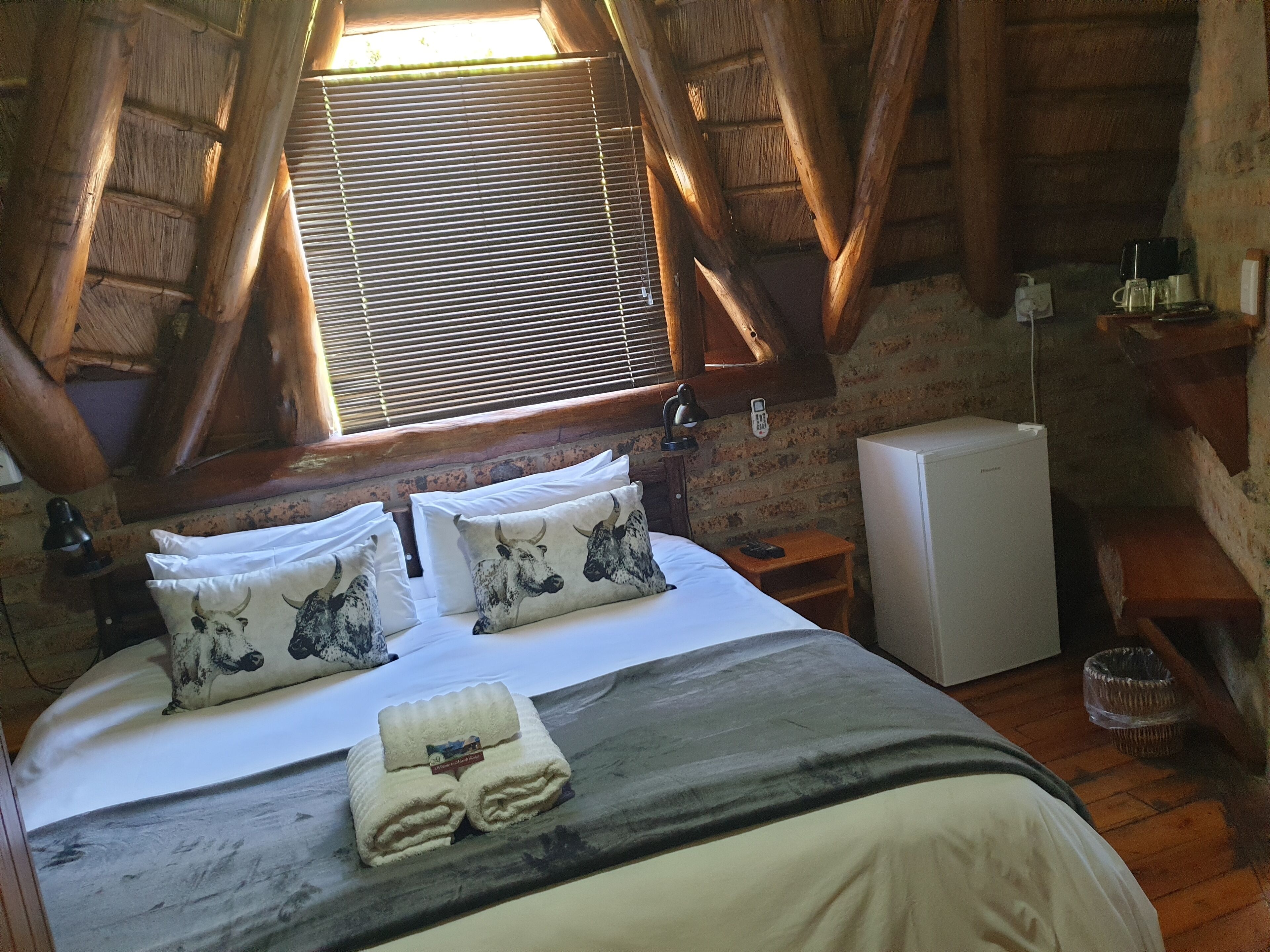 A cozy room with a wooden ceiling, a bed with white bedding and two decorative pillows, a small bedside table with a lamp, and a refrigerator.