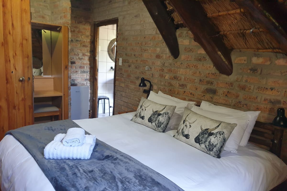 A bedroom with a large bed, a wooden wardrobe, a mirror, and a brick wall.