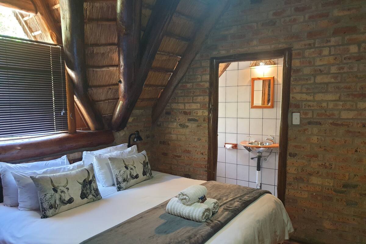 A bedroom with a bed, pillows, a bedside lamp, a bathroom with a sink and mirror, and a brick wall.