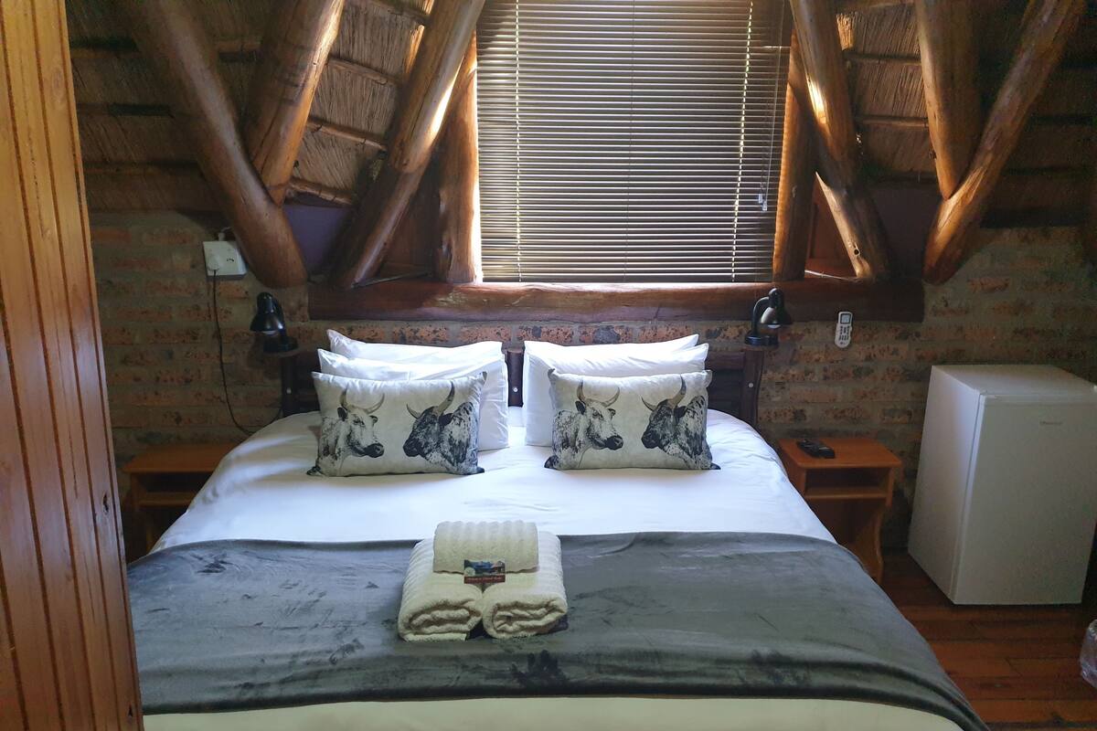 A bedroom with a loft-style ceiling, a bed with two decorative pillows, a bedside table with a lamp, and a small refrigerator.